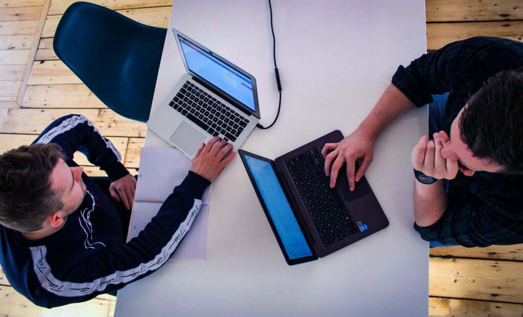 Zwei Personen an weißem Tisch mit Laptops, eine schreibt, eine tippt; Ansicht von oben.