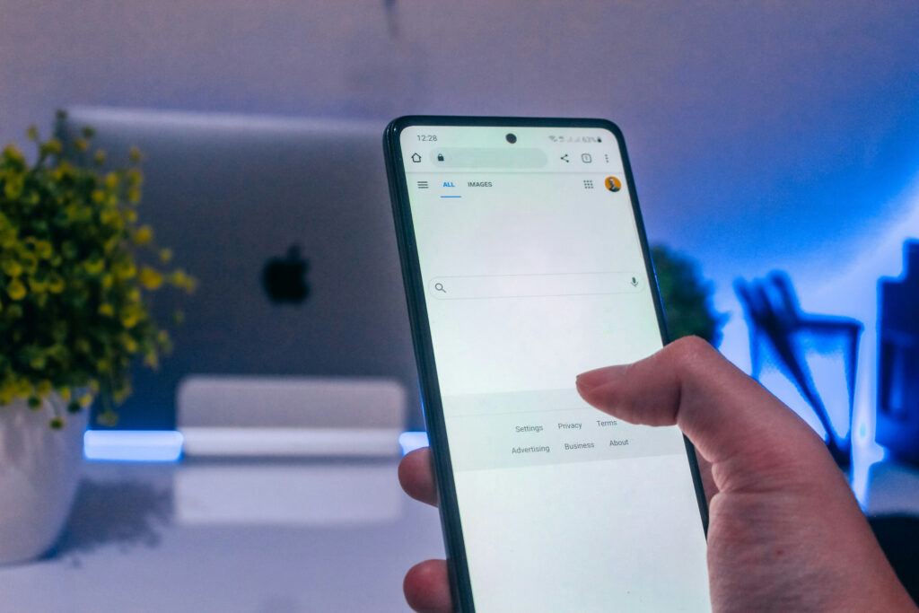 Person hält Smartphone mit Google-Suche; im Hintergrund Schreibtisch mit PC, Pflanze, Stuhl.