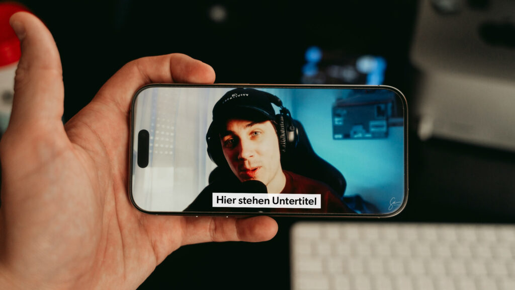 Hand hält Smartphone mit Video und Untertitel „Hier stehen Untertitel“, Tastatur im Hintergrund.