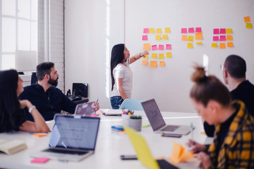 Person präsentiert am Whiteboard mit Haftnotizen; vier Personen mit Laptops hören im Büro zu.