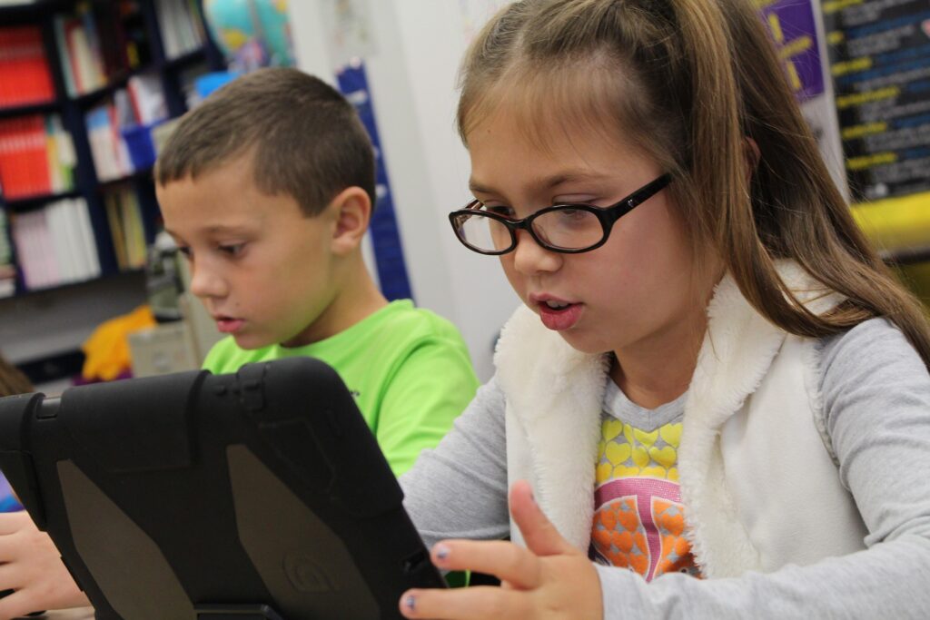 Zwei Kinder im Klassenzimmer schauen auf ein Tablet; Mädchen mit Brille, Junge im grünen Hemd.