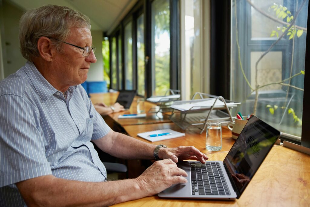 Älterer Mann mit Brille tippt am Laptop; im Hintergrund Papiere und eine weitere Person.